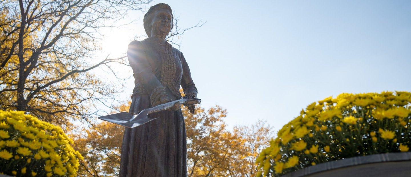 Bronze statue outdoors of a woman in historic clothing, holding a shovel.